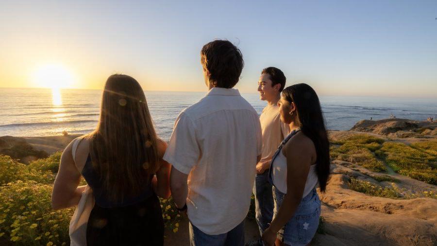 Four students stand on Sunset Cliffs together. They are smiling and looking at the sunset in the horizon.